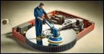 A surreal scene showing a standing maintenance person using a floor polisher on a giant hard disk platter. The maintenance worker, dressed in a standard blue jumpsuit and safety shoes, is actively operating the large, mechanical floor polisher.