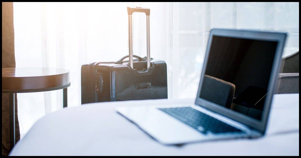 Laptop on hotel bed with suitcase in the background.