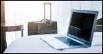 Laptop on hotel bed with suitcase in the background.